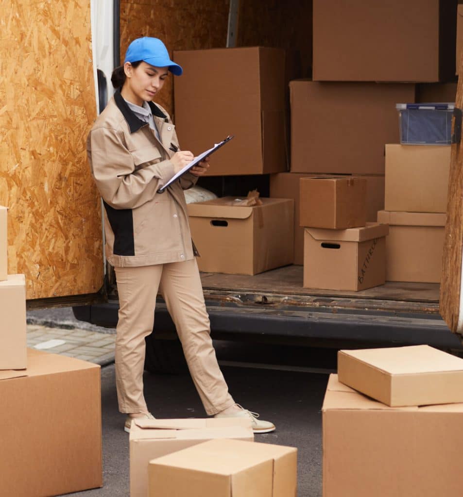 A delivery personnel in a beige uniform and blue cap stands beside a truck filled with cardboard boxes, taking notes on a clipboard.