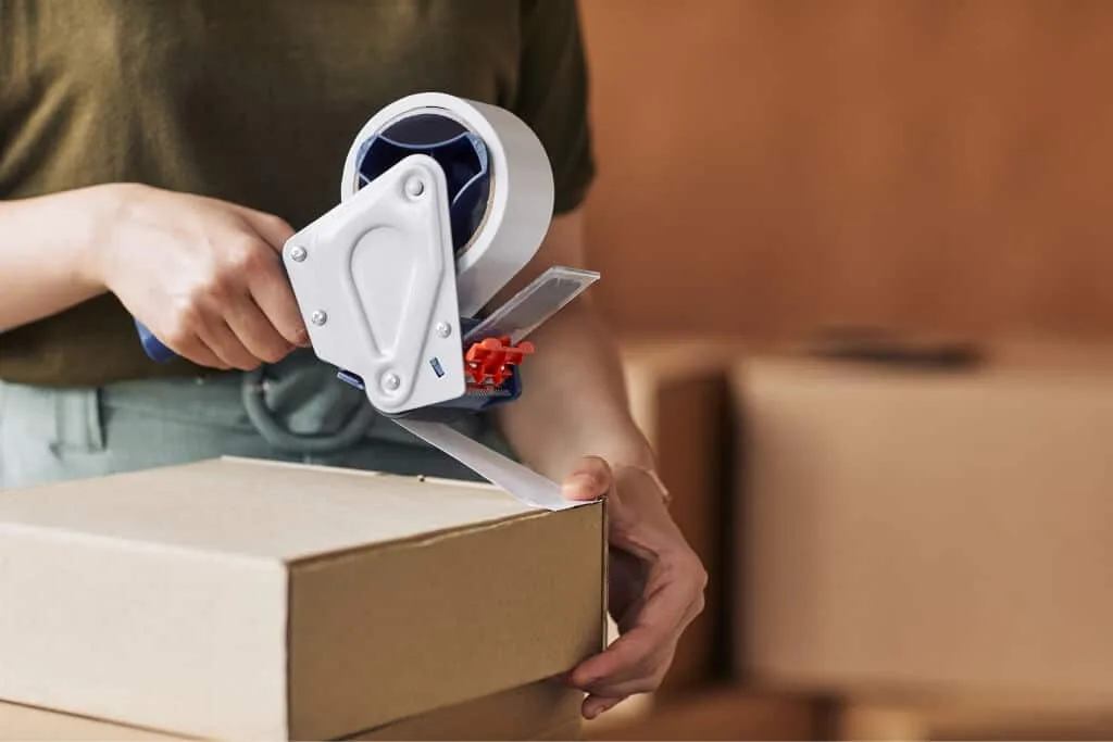 Individual using a tape dispenser to seal a cardboard box, showcasing packing supplies for shipping and storage.