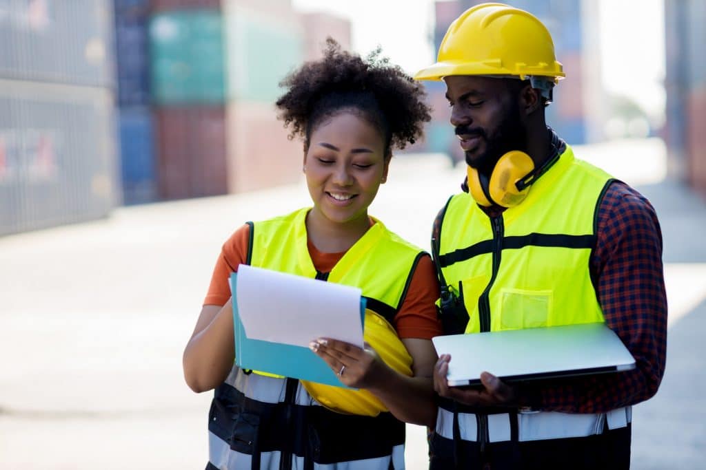 Two construction workers in safety vests and helmets reviewing documents and a laptop in a shipping yard.