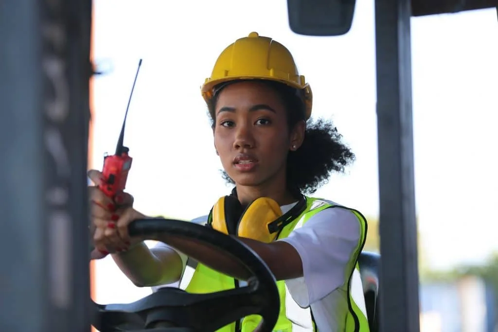 Female forklift operator wearing a yellow hard hat and safety vest, holding a radio while operating a forklift in a work environment.