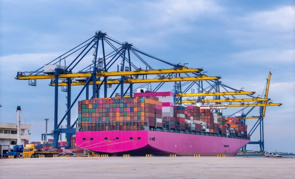 A large pink container ship docked at a port with numerous colorful shipping containers stacked on its deck and cranes in the background.